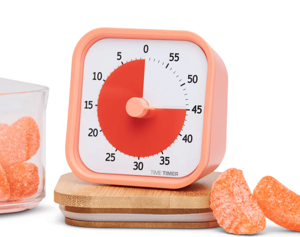 A close-up image of a visual timer set to 30 minutes, sitting on a bamboo coaster. Next to it, there is a glass container with orange gummy candy.